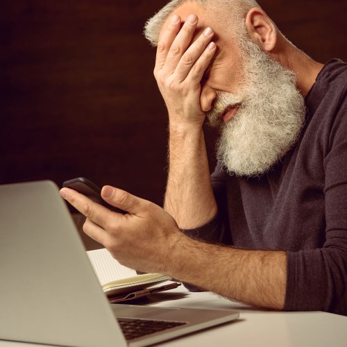 grey haired man sitting at table and holding smartphone with facepalm at home